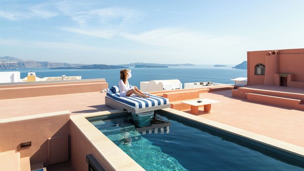 A person relaxing on a striped lounge chair by a pool with a view of the sea and distant landforms.