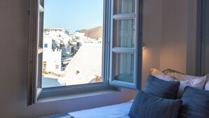 A room with a bed, a window with an open view of buildings, and a bedside table.