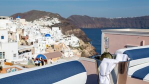 A balcony with a bottle of wine and a towel, overlooking a coastal town with white buildings and blue domes, and a view of the sea and mountains in the background.