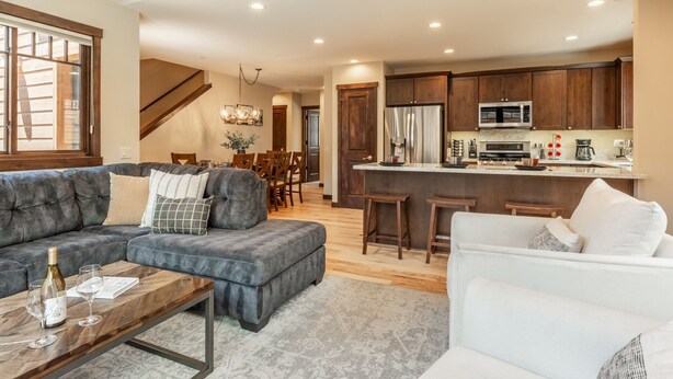 A modern living room with a grey sectional sofa, a white armchair, and a wooden coffee table. A kitchen with wooden cabinets and stainless steel appliances is visible in the background. - Hot Tub | Ping Pong Table | EV Charger (Mammoth Lakes)