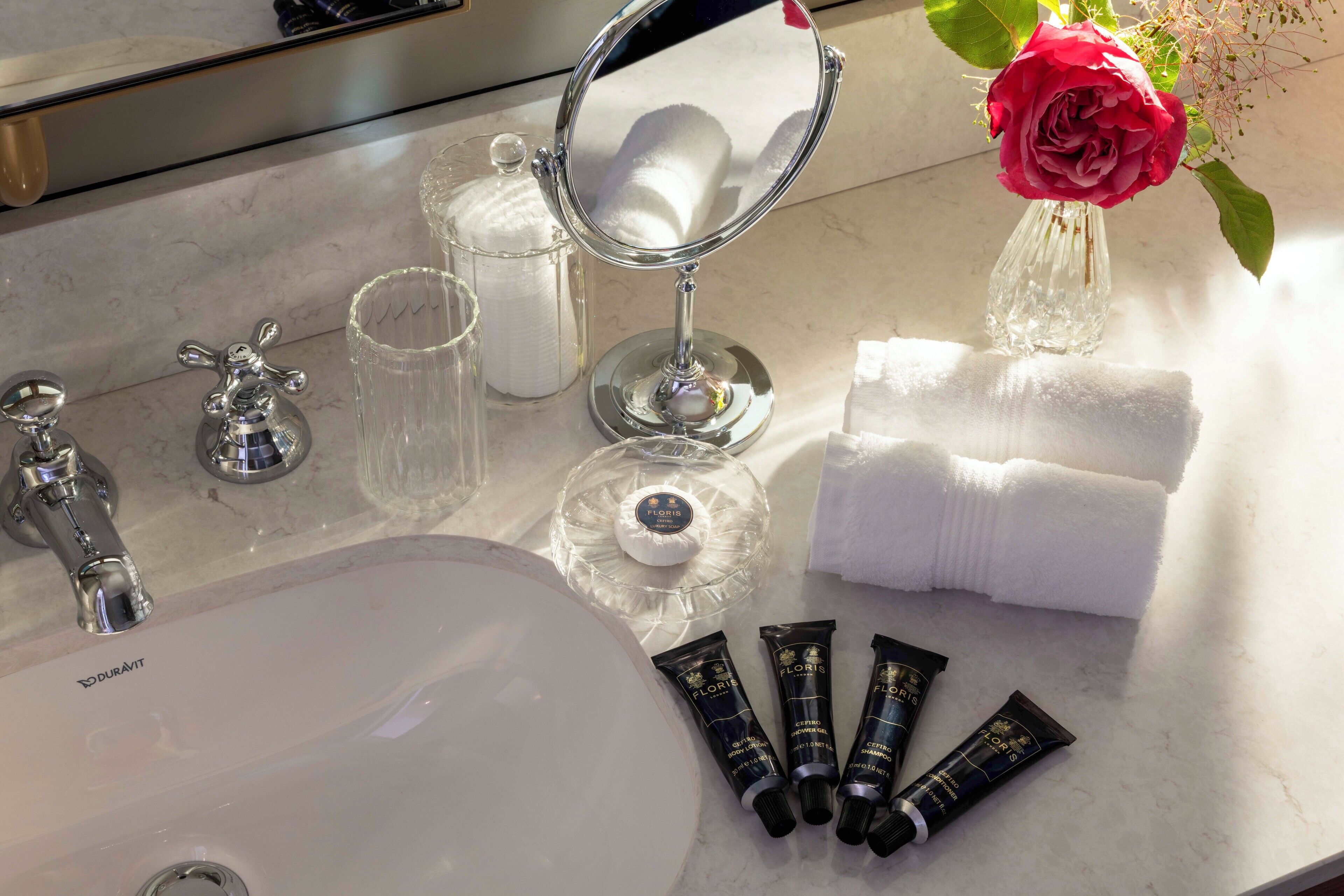 A bathroom vanity with a mirror, toiletries, and a vase with a rose.