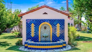 A small, tiled structure with a tiled archway and a fountain, surrounded by a well-maintained lawn and palm trees.