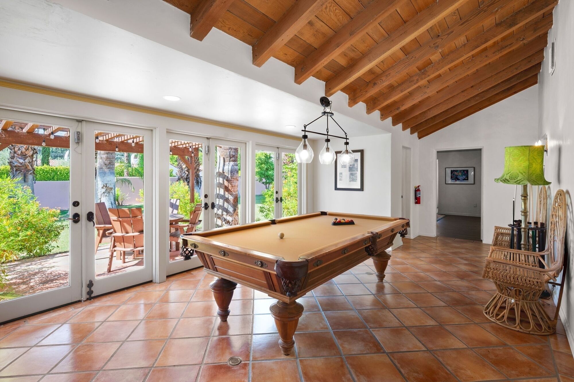 Une table de billard dans une pièce dotée d’un plafond en bois et d’un sol carrelé, de grandes fenêtres et d’un fauteuil en osier.