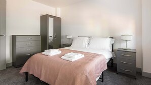 A neatly made bed with a pink bedspread and two towels, a dark dresser with drawers, a tall mirror, and two bedside tables with lamps. - Spacious & Bright 1 Bed Flat in Central Doncaster (South Yorkshire)