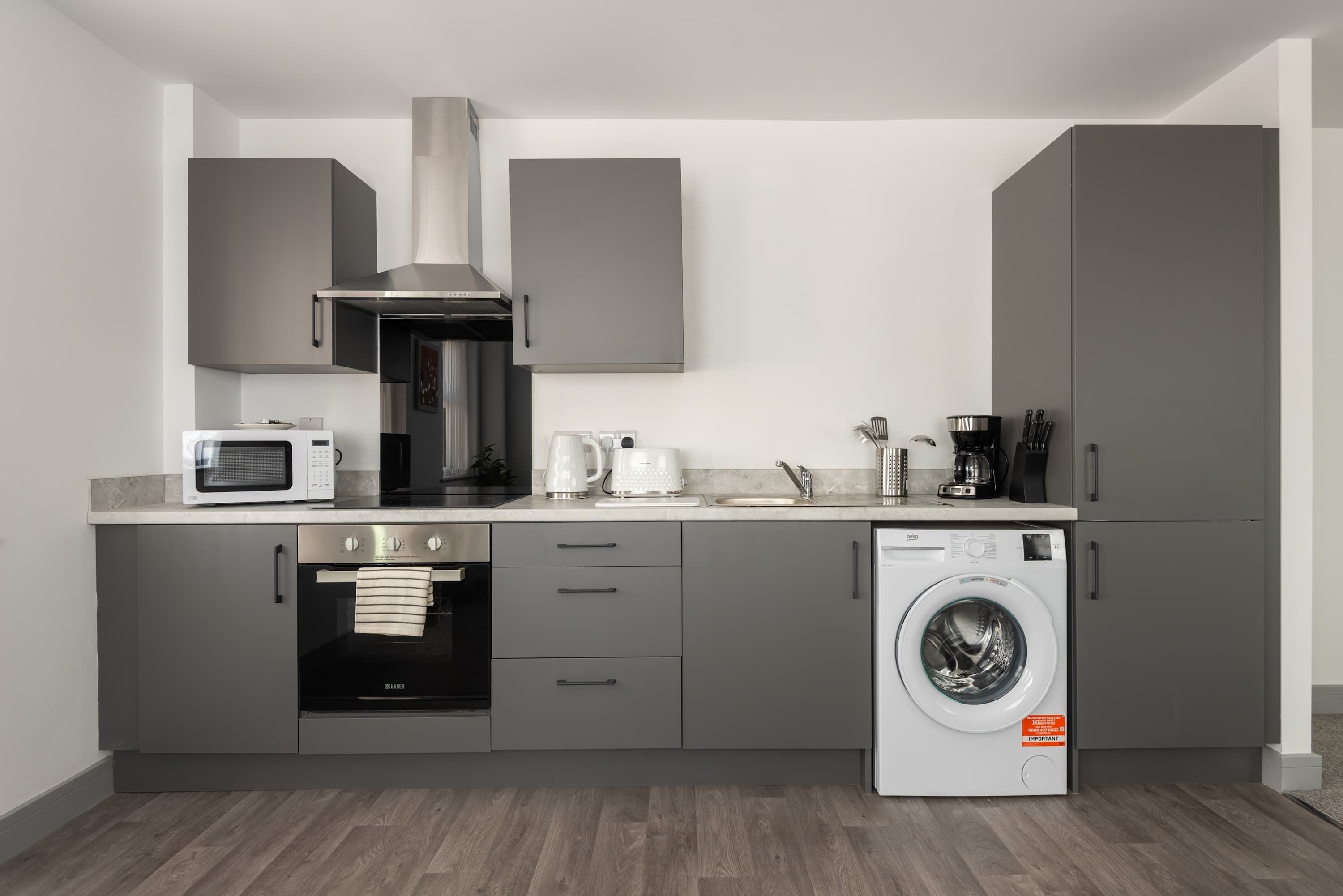 A modern kitchen with grey cabinets, a built-in oven, a microwave, a coffee maker, and a washing machine.
