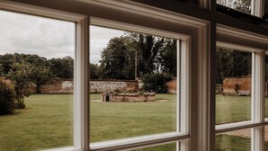 Uitzicht door een raam op een grasrijk erf met een bakstenen muur en bomen.