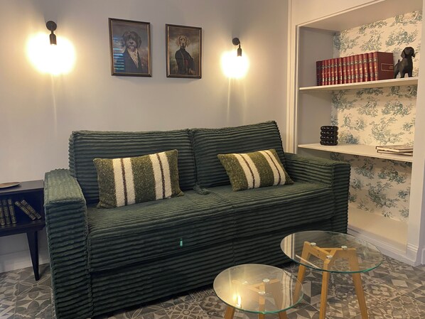 Un sofá verde con cojines a rayas, una mesa de centro de cristal y una estantería con libros y adornos.