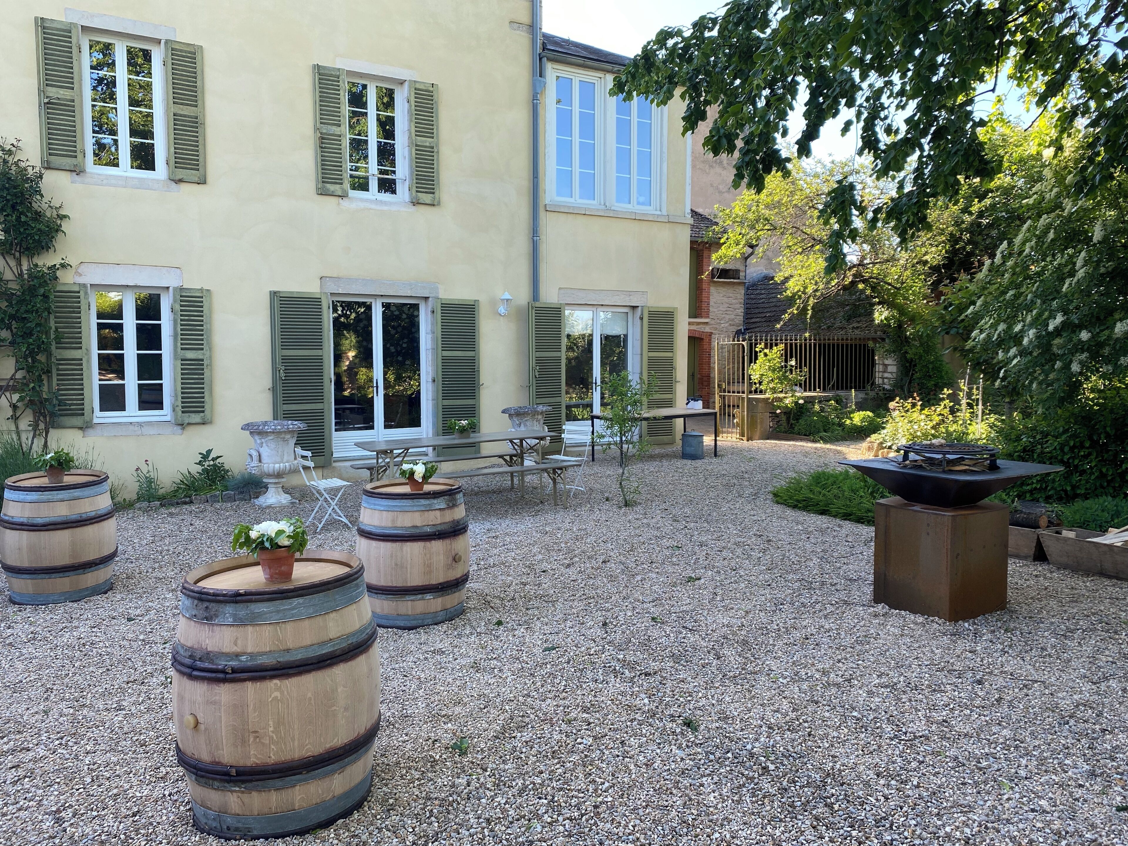 Une cour avec des tables et des chaises à l’extérieur, des tonneaux en bois et un bâtiment aux volets verts.