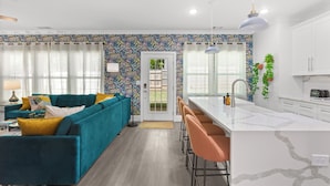 A modern living room with a teal sofa, a kitchen with white cabinets, and a bar area with orange stools.
