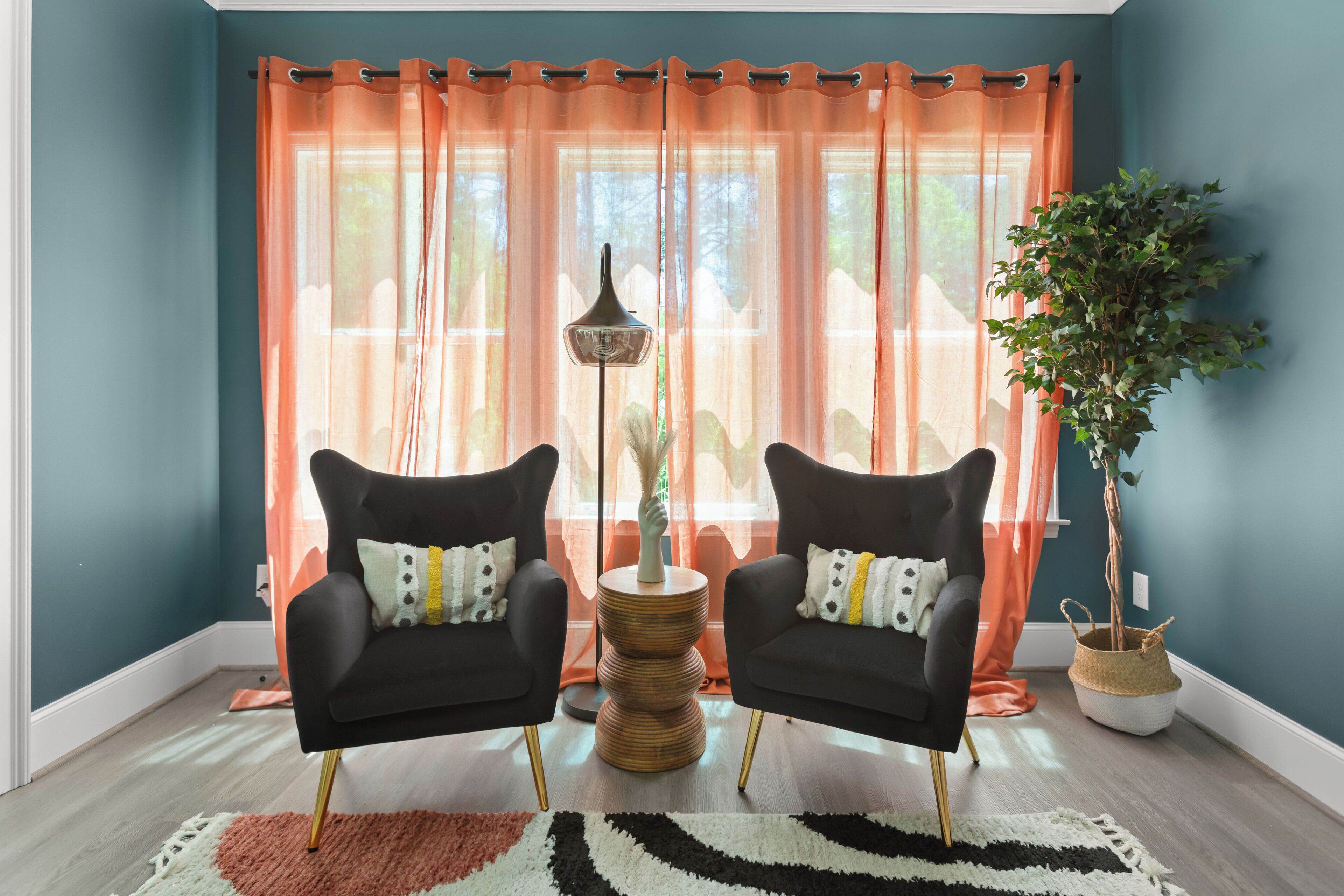A room with teal walls, two black armchairs with patterned cushions, a woven side table, a floor lamp, a large window with orange curtains, and a potted plant.
