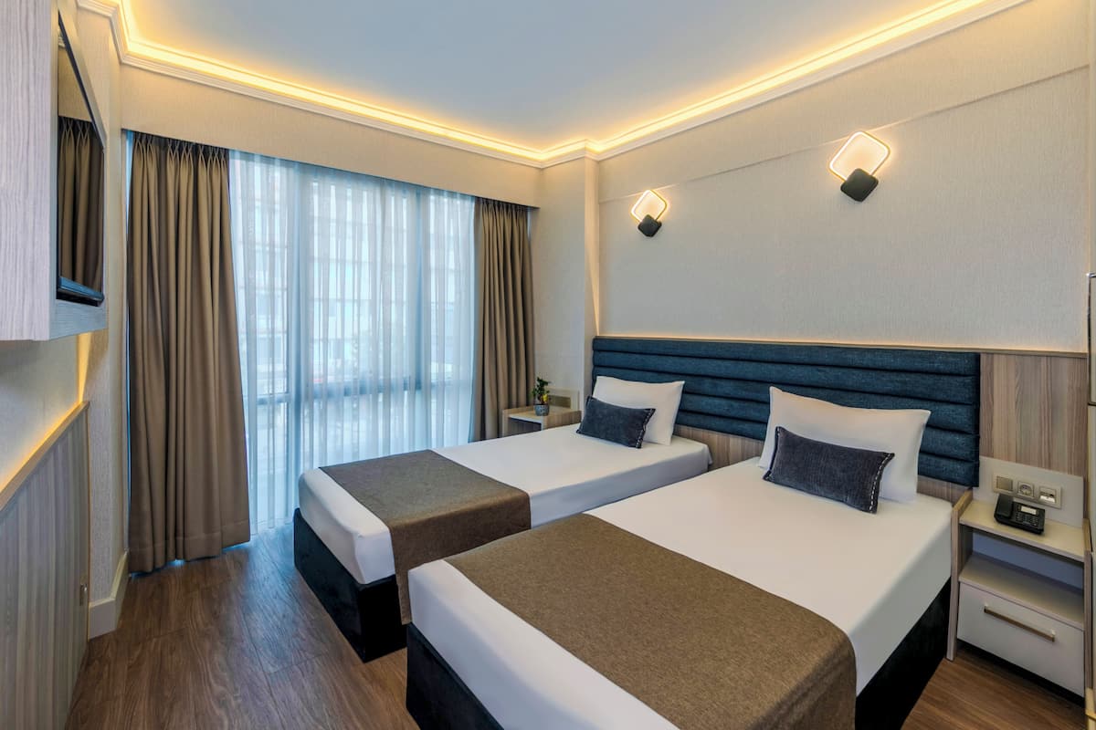 Habitación de hotel con dos camas, un ventanal con cortinas, una mesita de noche con lámpara y un televisor de pared.