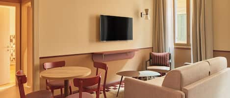 A hotel room with a sofa, a TV mounted on the wall, a dining table with chairs, and a small side table.