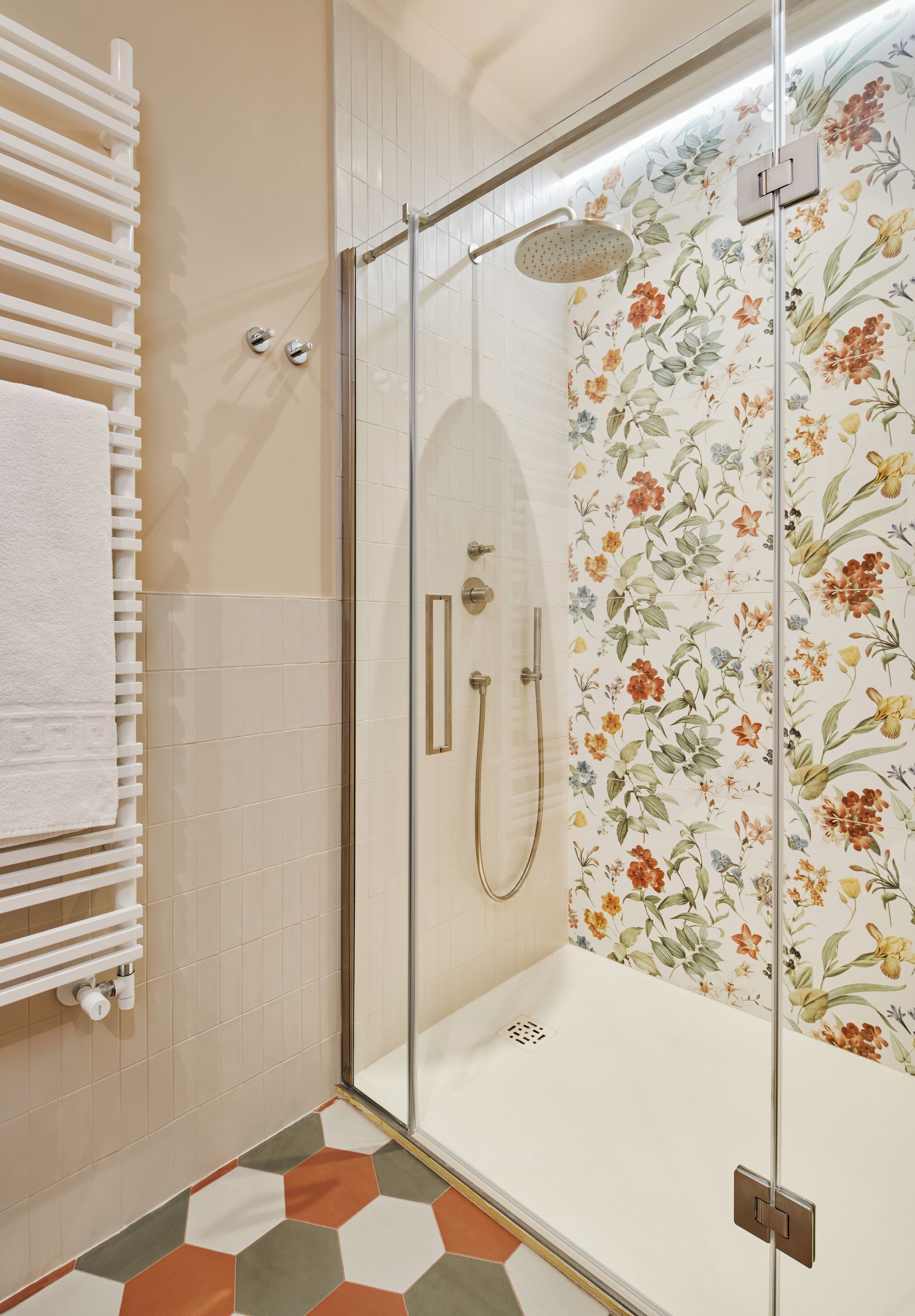 Une douche avec une porte en verre, un porte-serviettes blanc et un mur de douche orné d’un motif floral.