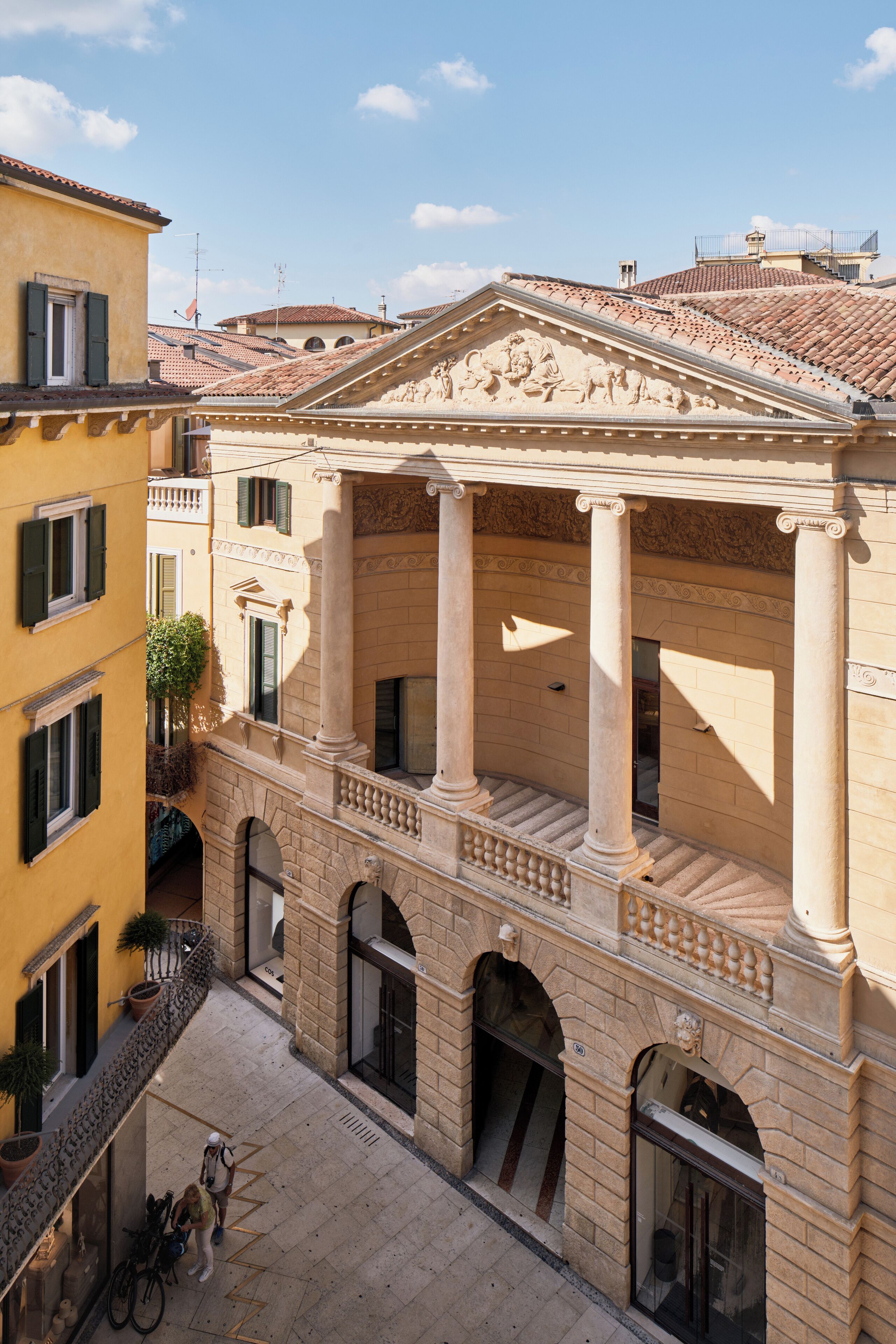 A courtyard with a building featuring classical columns, arched doorways, and a tiled roof.