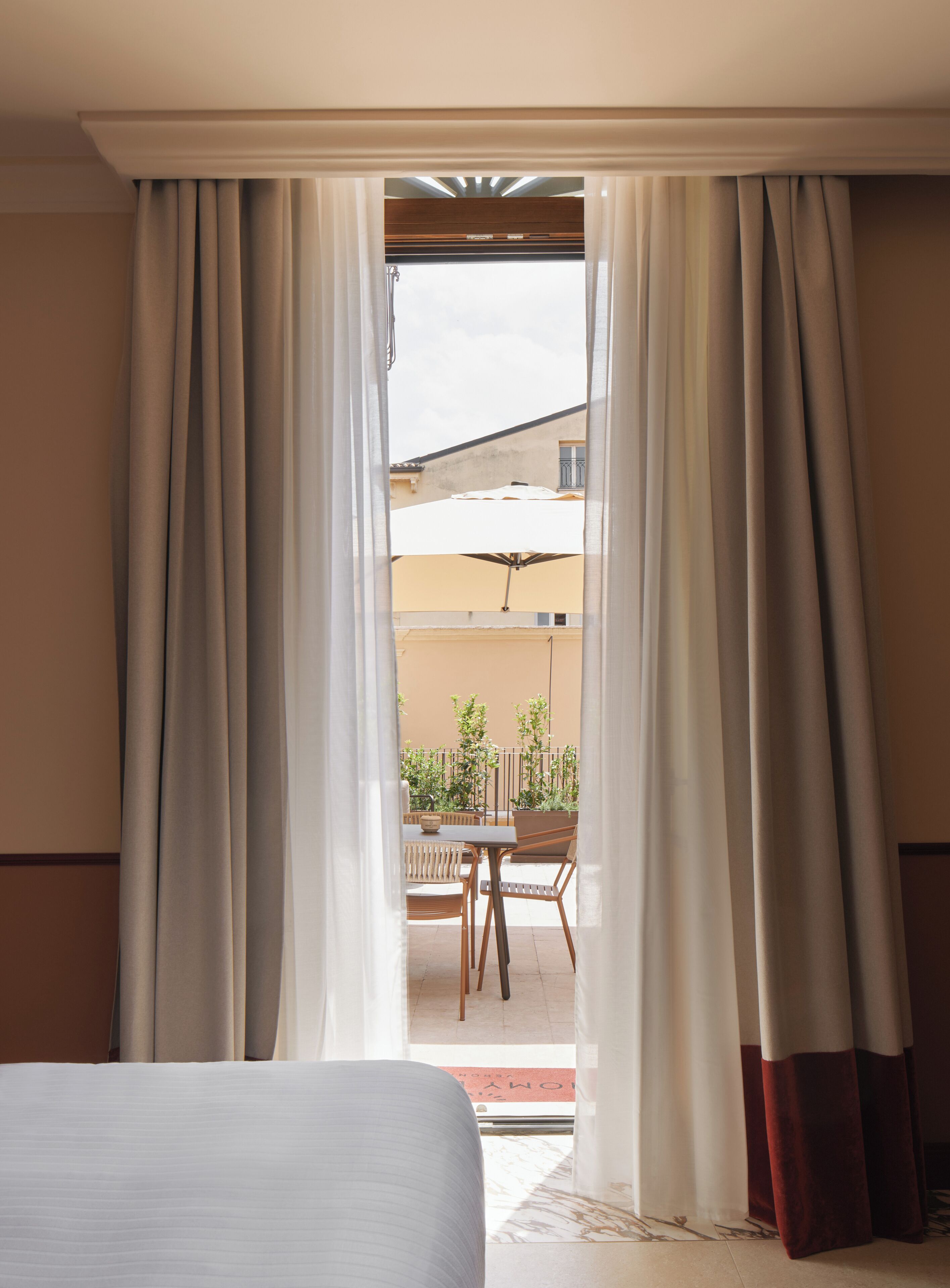 Habitación de hotel con cama, cortinas y puerta abierta que da a un espacio exterior con una mesa y sillas.