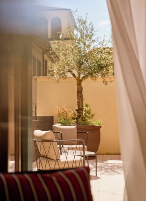 A sunlit outdoor seating area with a table and chairs, a large potted plant, and a building with arched windows in the background.
