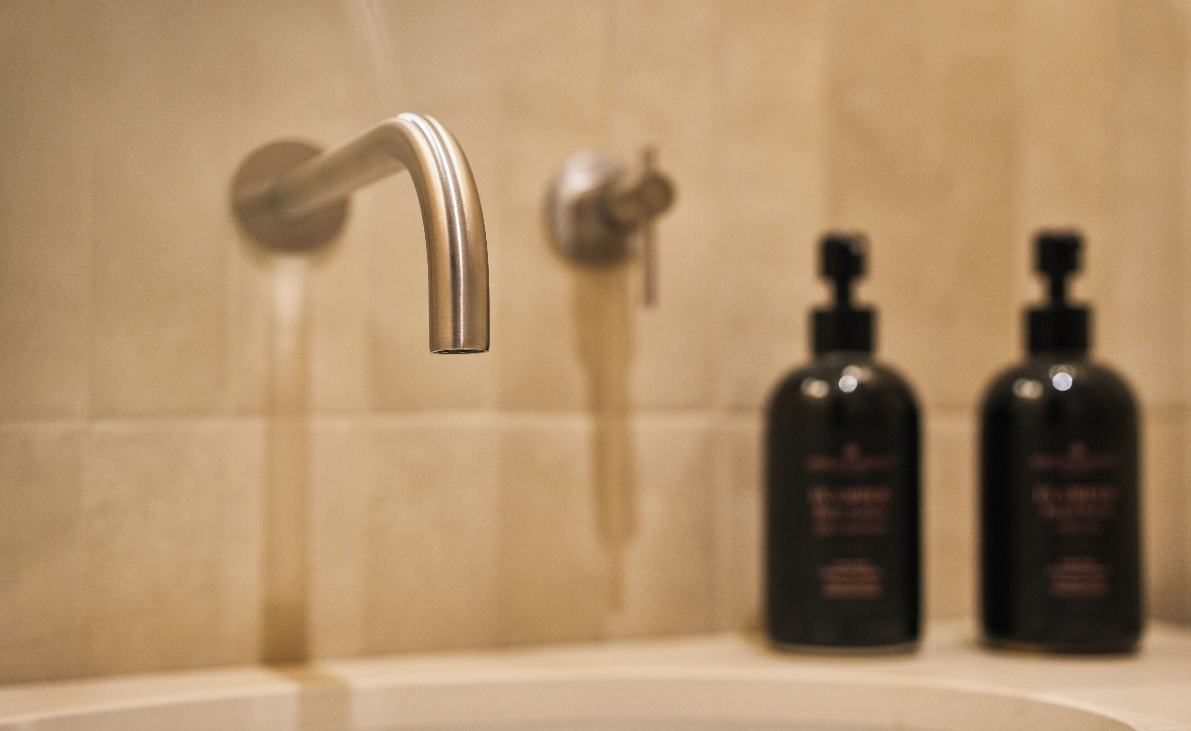 A bathroom sink with a chrome faucet, two black soap dispensers, and a tiled wall.