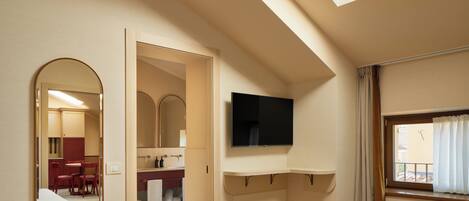 Un dormitorio con tragaluz, televisor montado en la pared, cama, espejo y un baño visible a través de una puerta abierta.