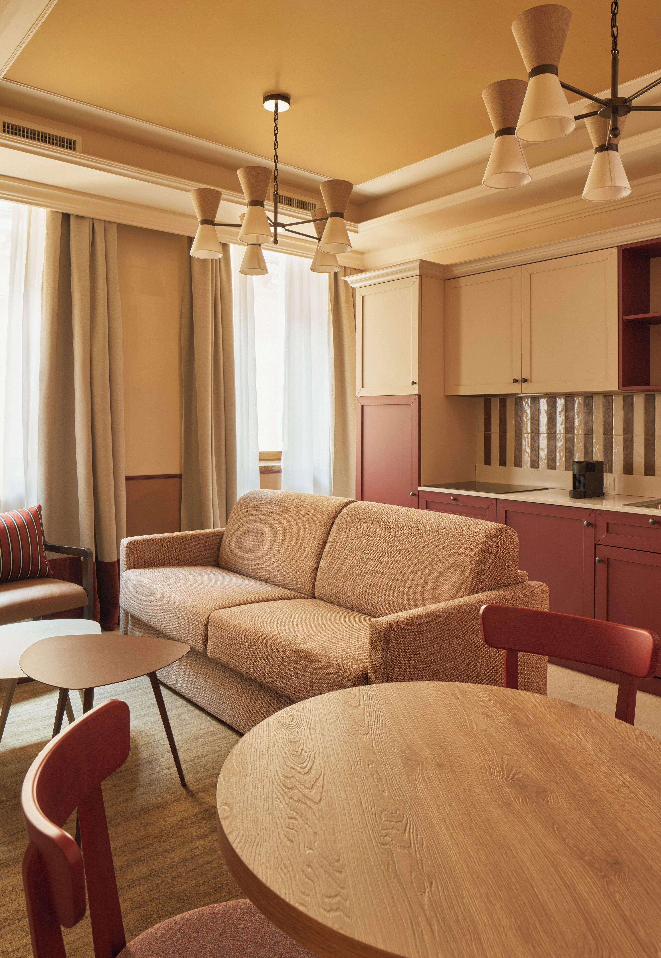 Una sala de estar moderna con un sofá beige, una mesa de centro de madera y un sillón rojo. Se ve un área de cocina con armarios y una encimera al fondo.