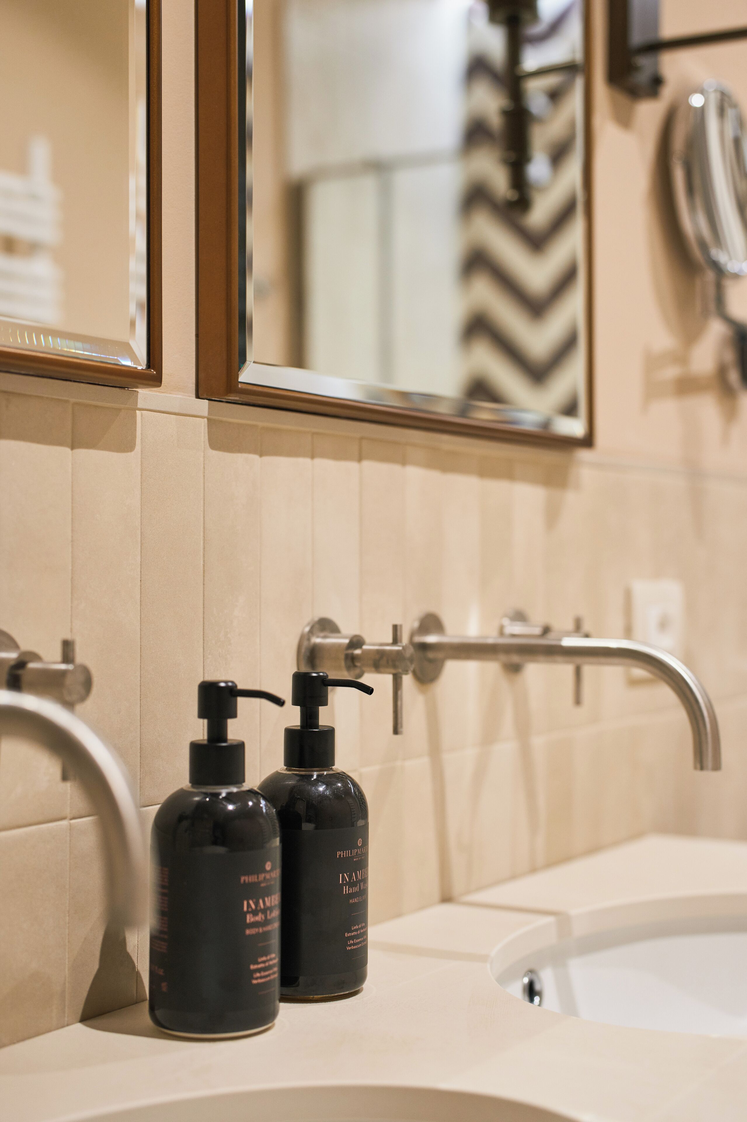 Une salle de bain avec un mur recouvert de carreaux beiges, un lavabo blanc, deux distributeurs de savon noir et un robinet chromé.