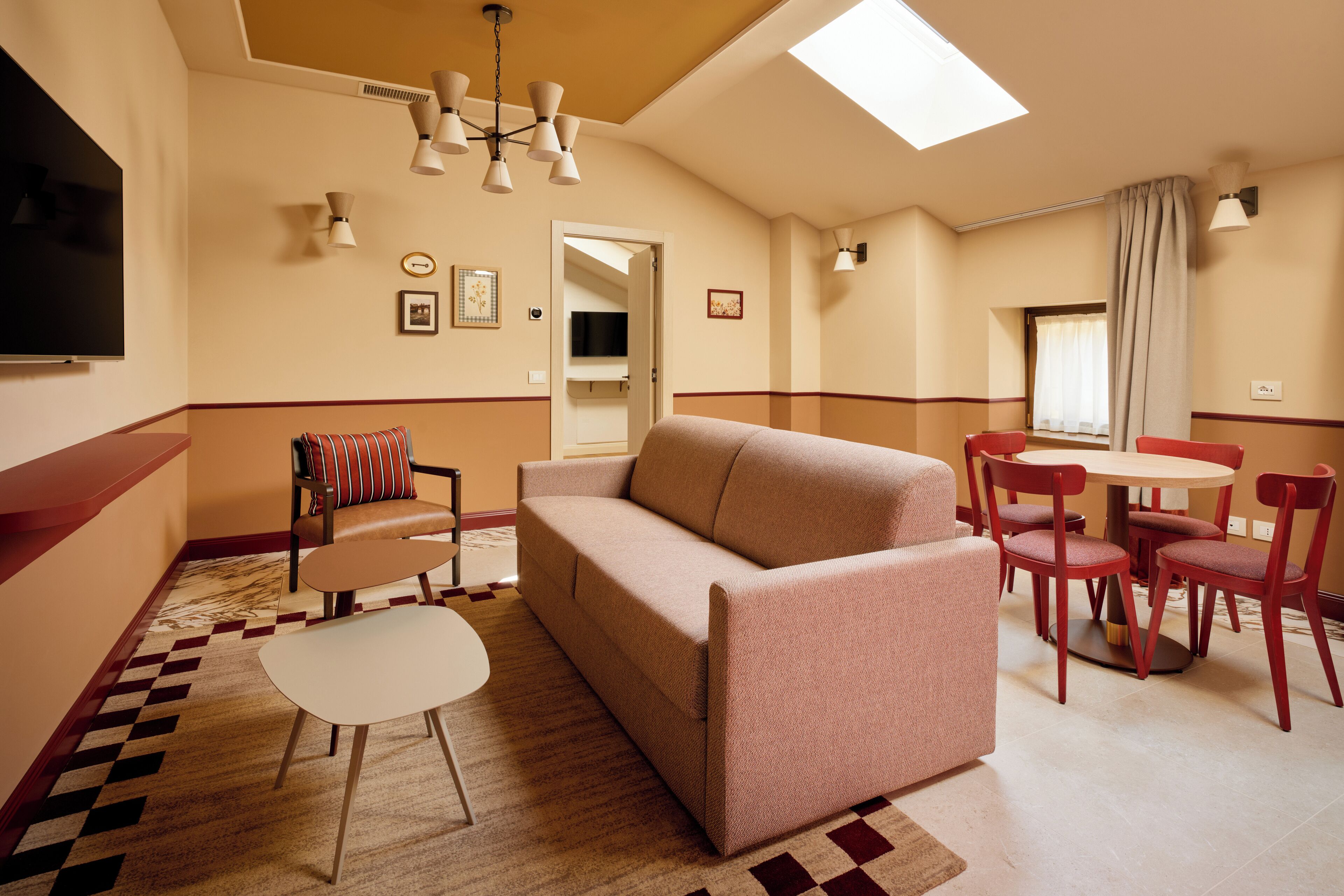 A modern living room with a sofa, chairs, a small table, and a TV. There is a dining area with a table and chairs, and a window with curtains.