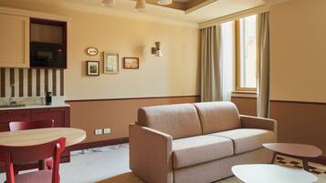 A modern hotel room with a sofa, a dining table, chairs, a small side table, a microwave, and a wall-mounted light fixture.