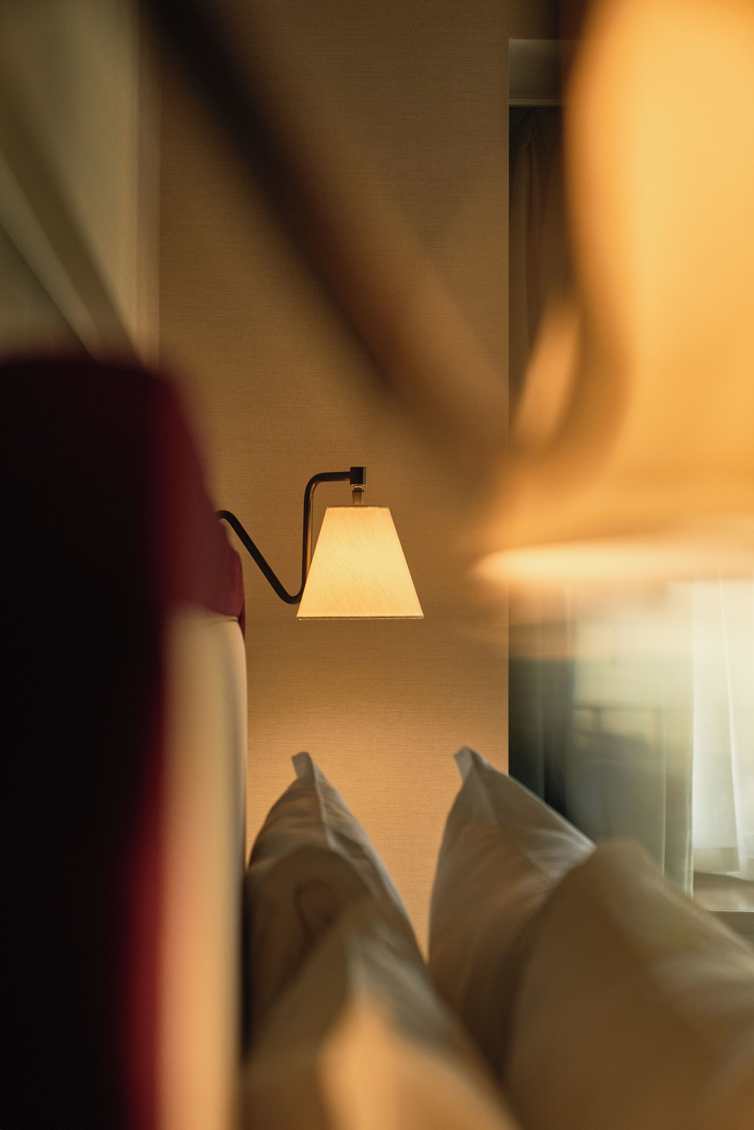 Quarto com luminária de cabeceira, janela com cortinas e cama com almofadas.