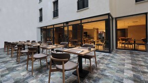 Un área de comedor al aire libre con varias mesas y sillas, piso de baldosas y un edificio con amplias ventanas y puertas.