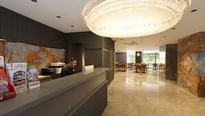 Zona de recepción de hotel con una lámpara de araña moderna, un mostrador con folletos y un comedor con mesas y sillas.