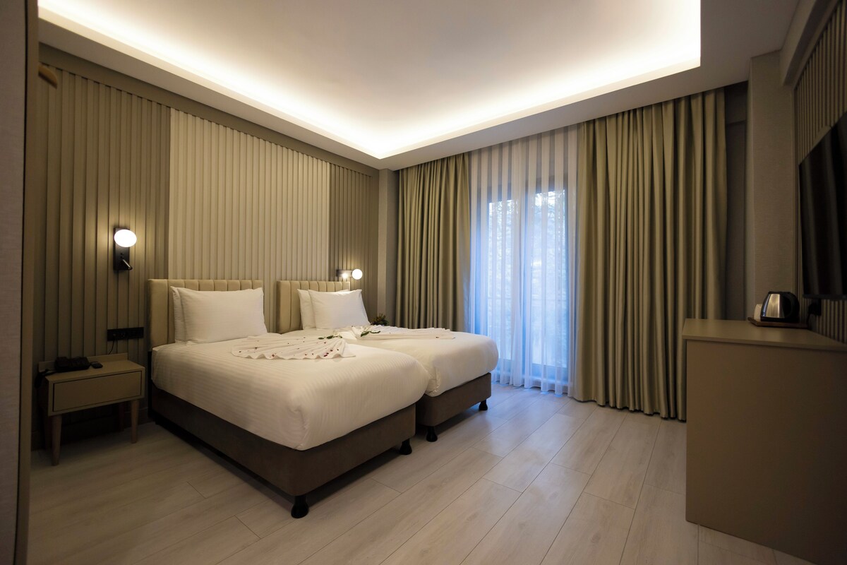 A hotel room with two beds, a bedside table with a lamp, a desk, and a large window with curtains.