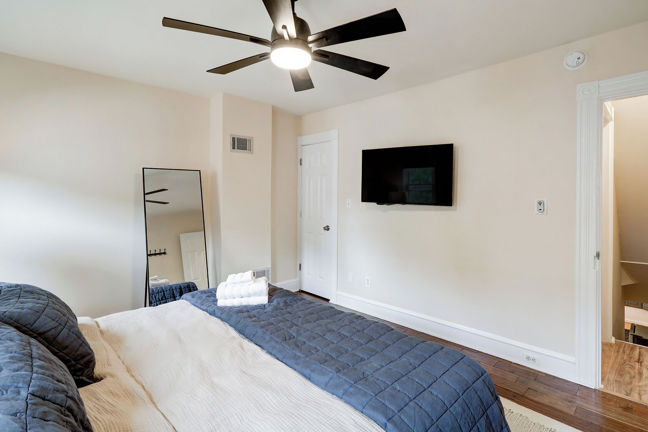 A bedroom with a bed, a ceiling fan, a TV mounted on the wall, and a door leading to another room.