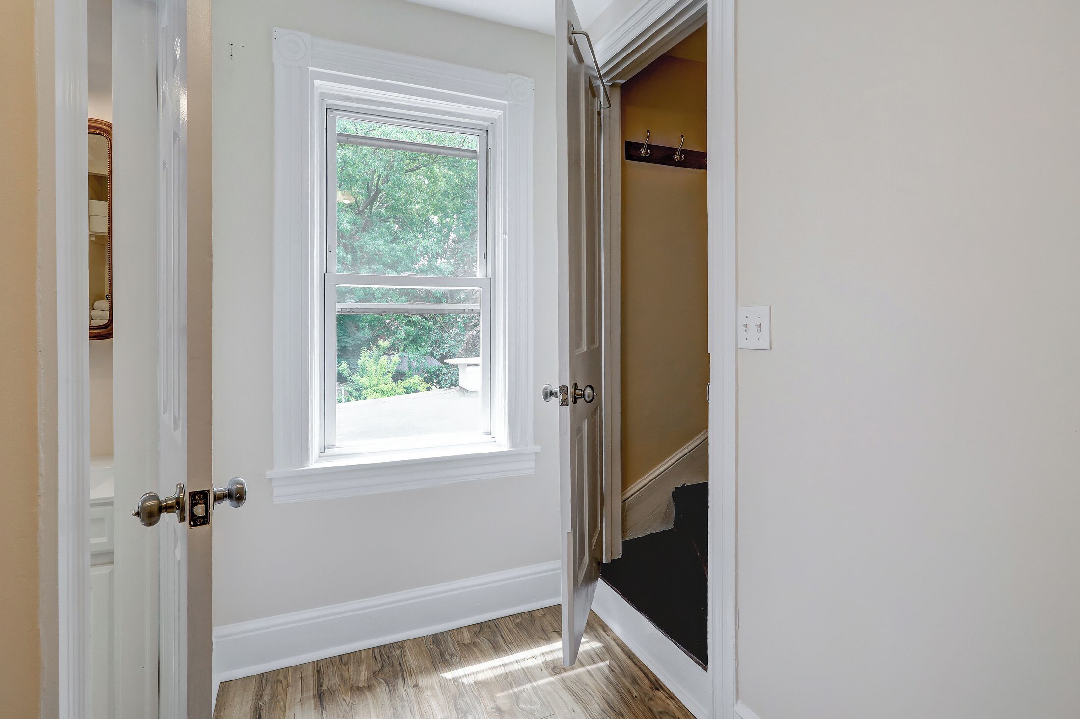 A room with a white door, a window with a view of greenery, and a coat rack on the wall.