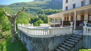 A stone staircase leading to a balcony with a white balustrade, surrounded by lush greenery and a mountainous backdrop.