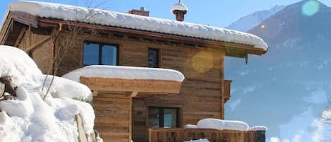 A wooden chalet with a snow-covered roof, a wooden porch, and a mountain backdrop.