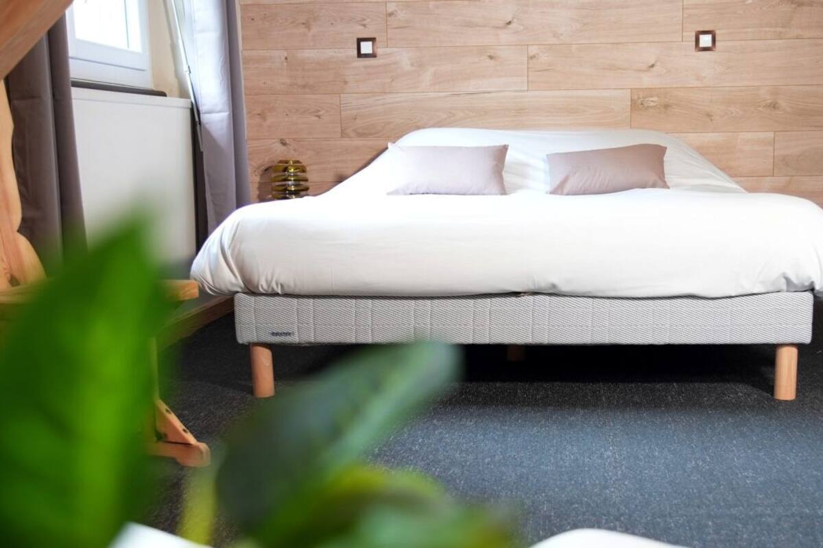 Cama com colchão cinza e roupa de cama branca em quarto com paredes de madeira e carpete cinza.