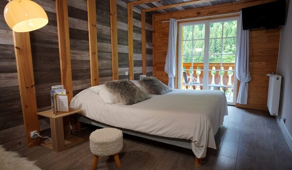 A wooden cabin bedroom with a bed, a nightstand, a small stool, and a window with a view of greenery. - La Sapiniere (Saint-Lary-Soulan)