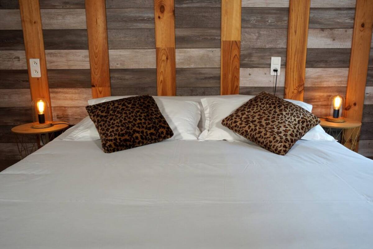 Cama com roupa branca e duas almofadas de padrão de leopardo, apoiada em parede de madeira com ripas verticais.