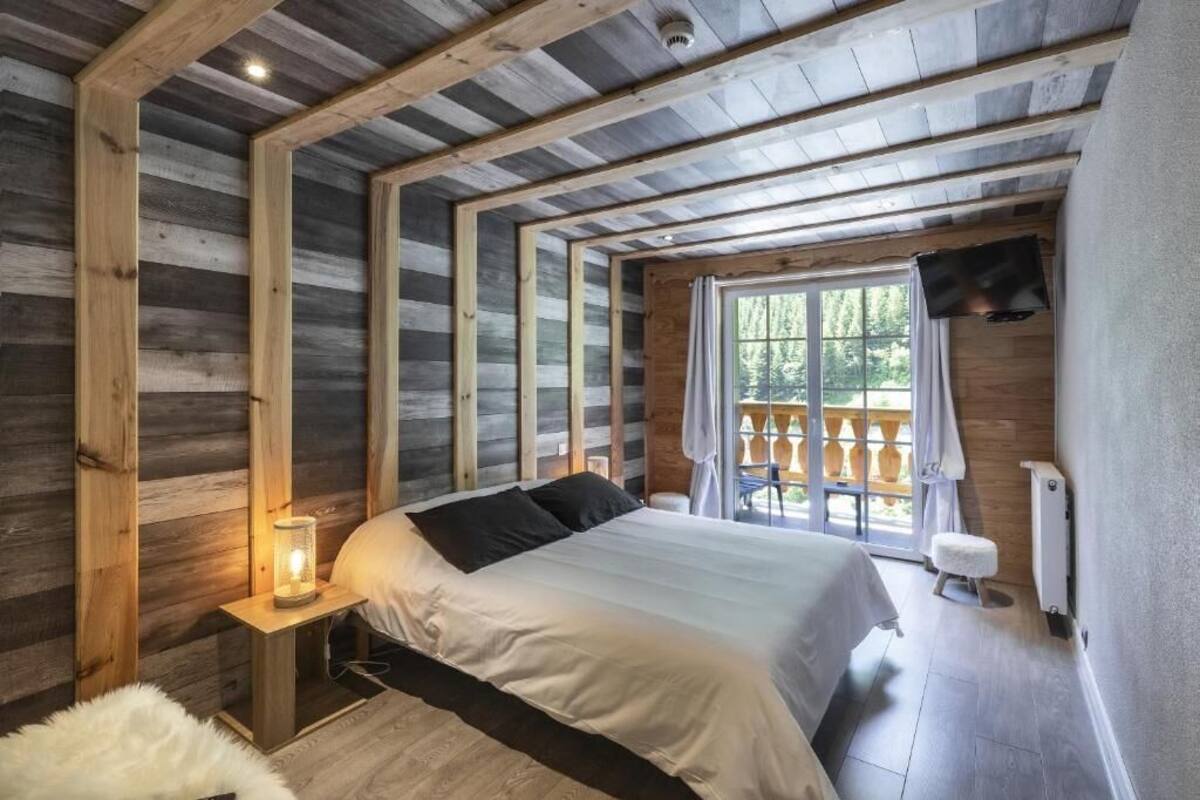 Quarto com teto de madeira, cama com roupa de cama branca, mesa de cabeceira com luminária, janela com cortinas e televisão fixada na parede.
