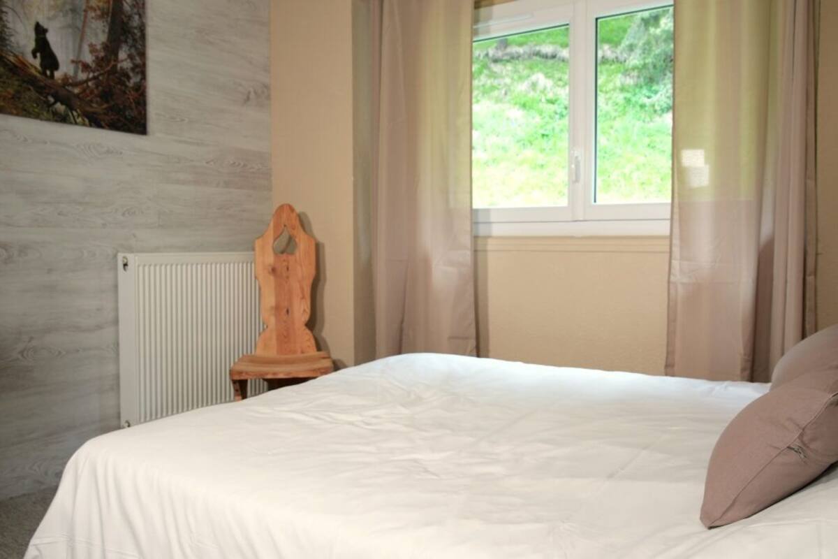 Um quarto com cama, uma cadeira de madeira, um radiador e uma janela com cortinas.