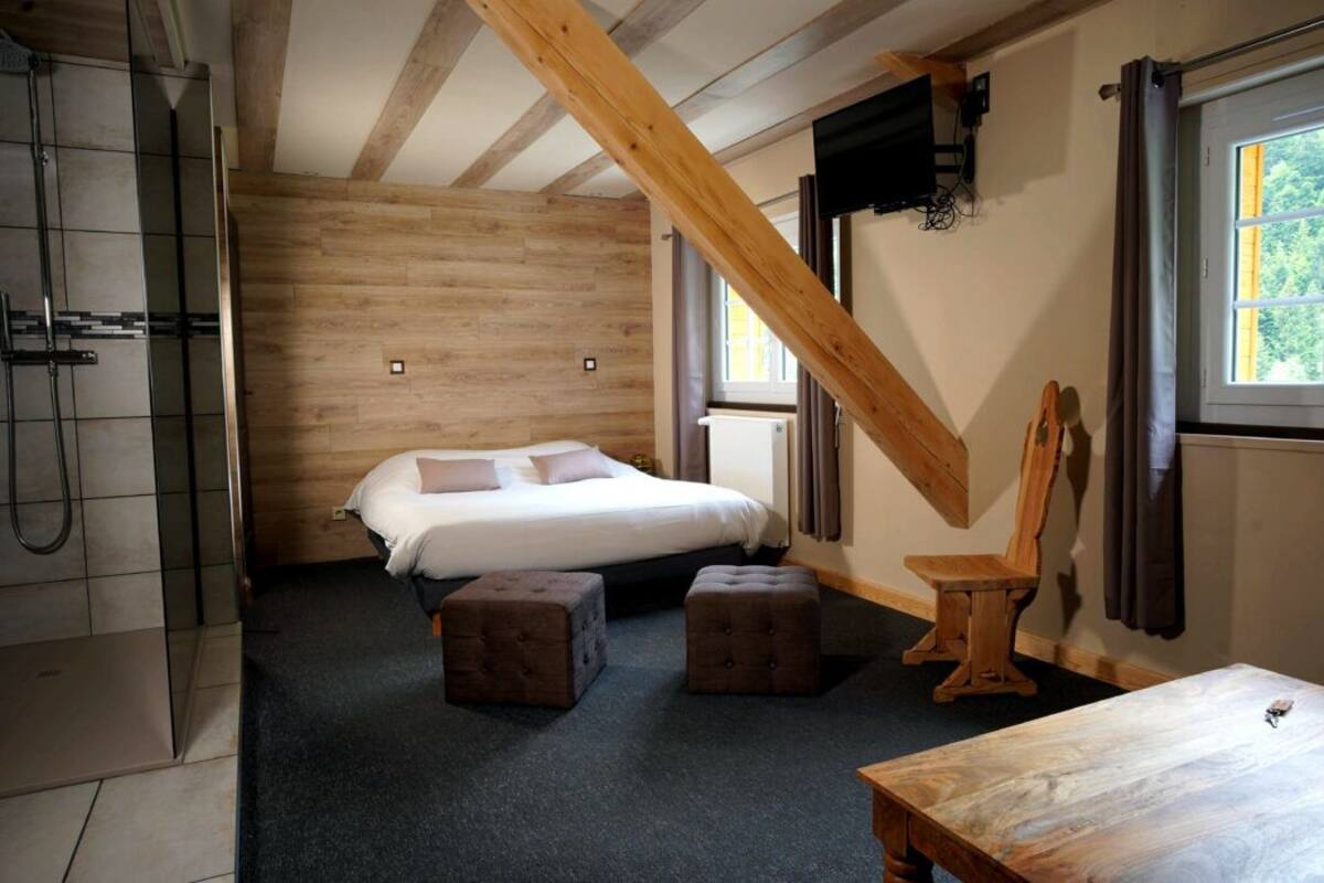 Quarto com cama, dois pufes, uma mesa de madeira, chuveiro e janela com vista para árvores.