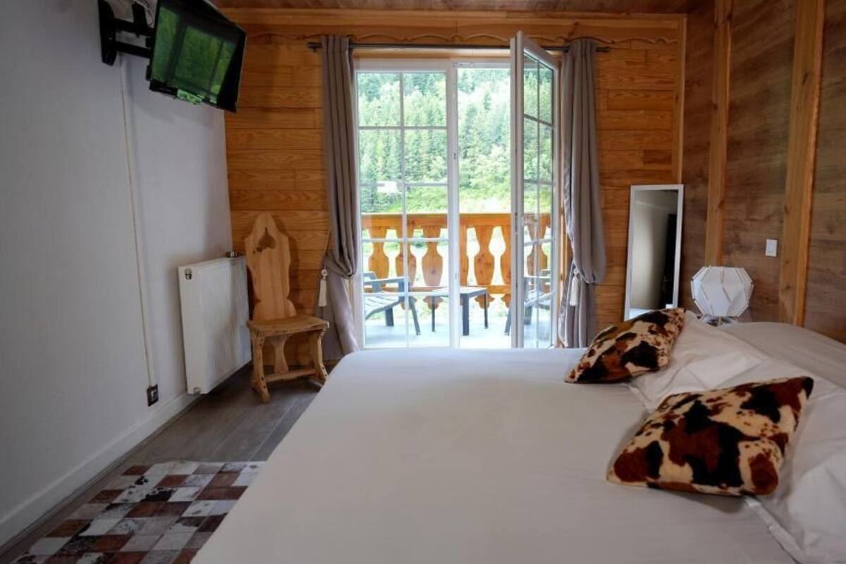 Quarto com uma cama grande, uma cadeira de madeira, uma televisão e uma varanda com vista para árvores.