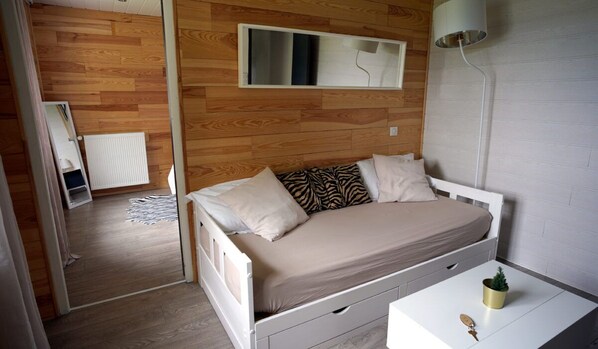 A bedroom with a wooden wall, a bed with a beige mattress and patterned pillows, a bedside table with a plant, and a white lamp. - La Sapiniere (Saint-Lary-Soulan)