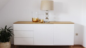 A white cabinet with a wooden top, two clear glasses with golden liquid, and a lamp with a beige shade. - 90 sqm apartment suitable for family (Bad Soden am Taunus)