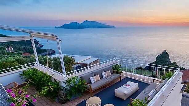 A rooftop terrace with a sofa, cushions, and a small table, overlooking a calm sea and distant mountains. - Casa Oscar - My Rental Homes (Marciano)