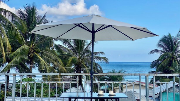 Ein Balkon mit Tisch und Sonnenschirm mit Blick auf einen Strand, Palmen und das Meer.