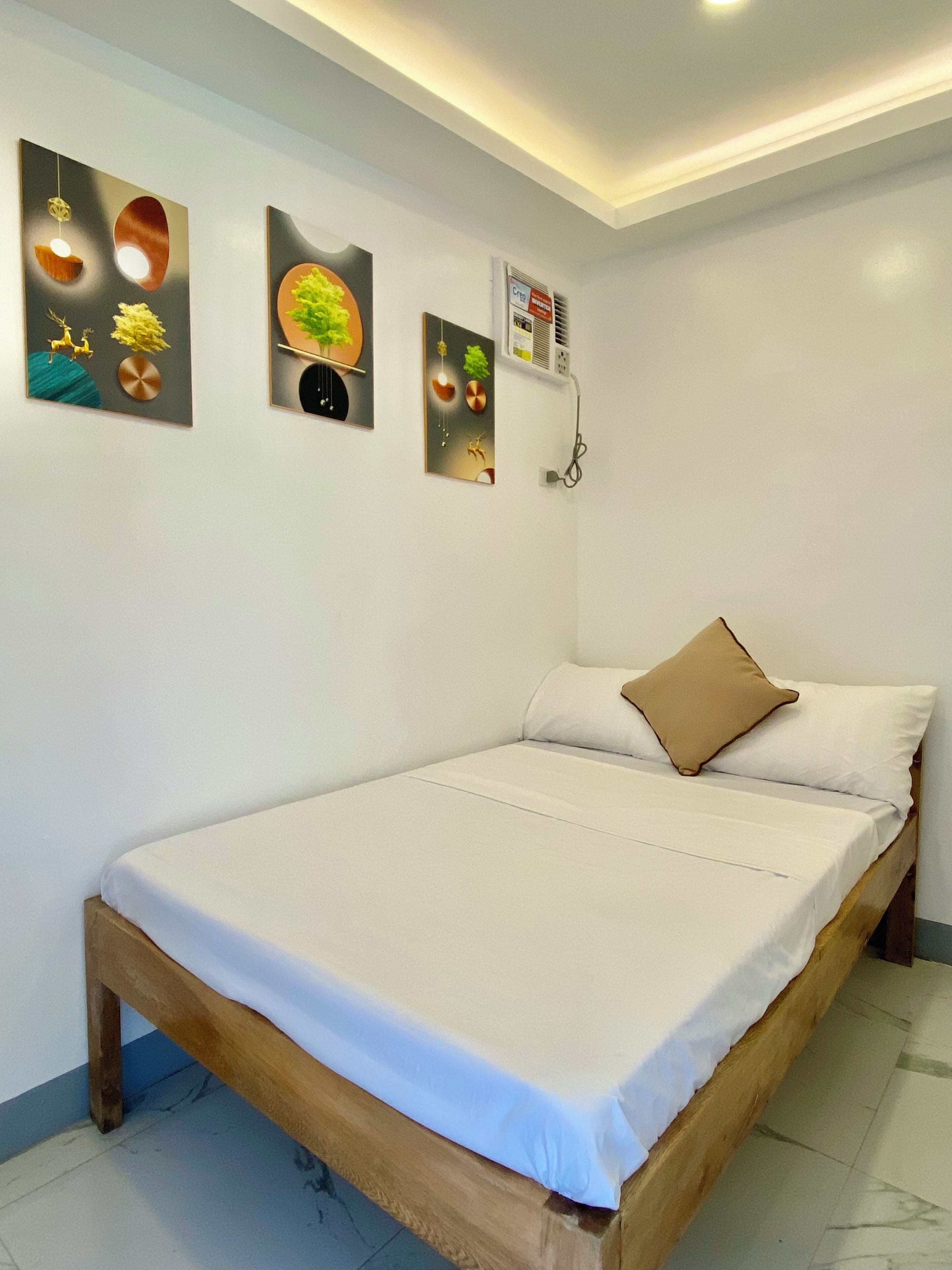 A neatly made bed with white linens and a single brown pillow, a wall-mounted air conditioner, and three framed artworks depicting abstract designs.