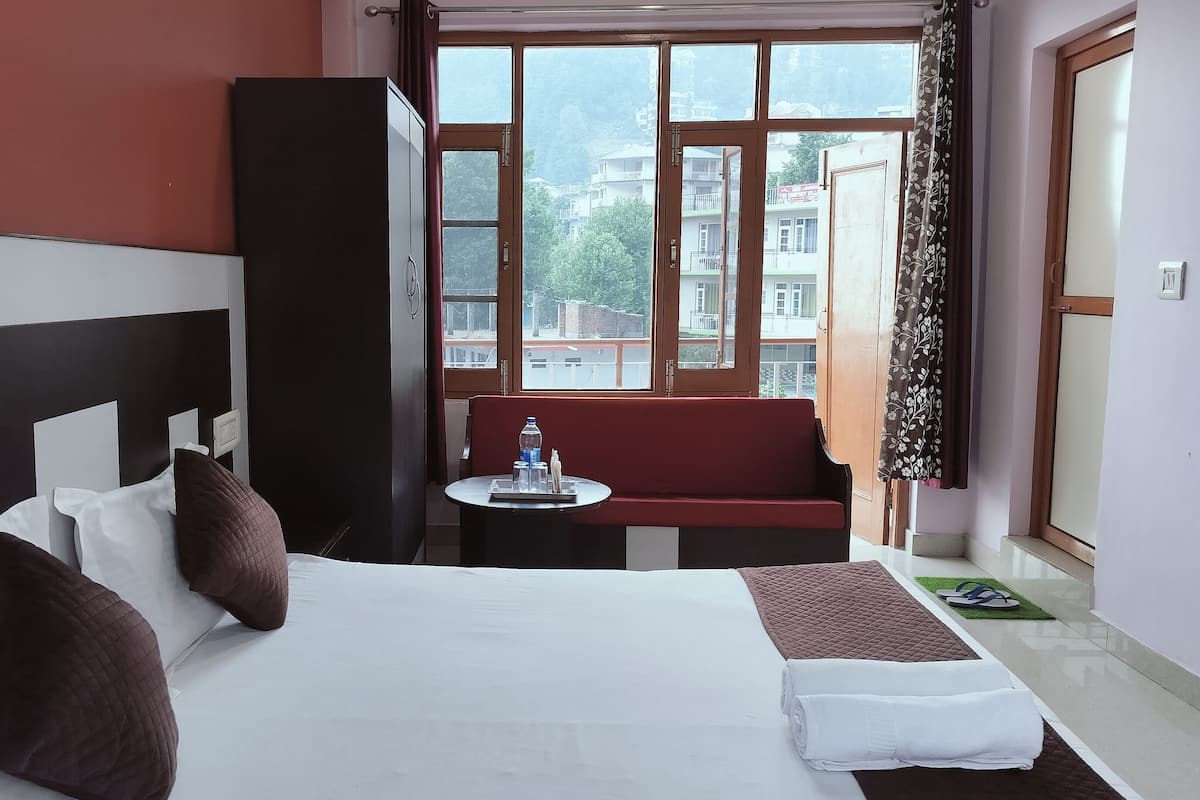 A hotel room with a bed, a small table with a water bottle, a red bench, and a view of buildings through large windows.