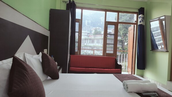 A hotel room with a bed, pillows, a window with a view of buildings, and a red bench by the window. - Hotel Adventure Inn 20 Metre from Swimming Pool (Dharamshala)