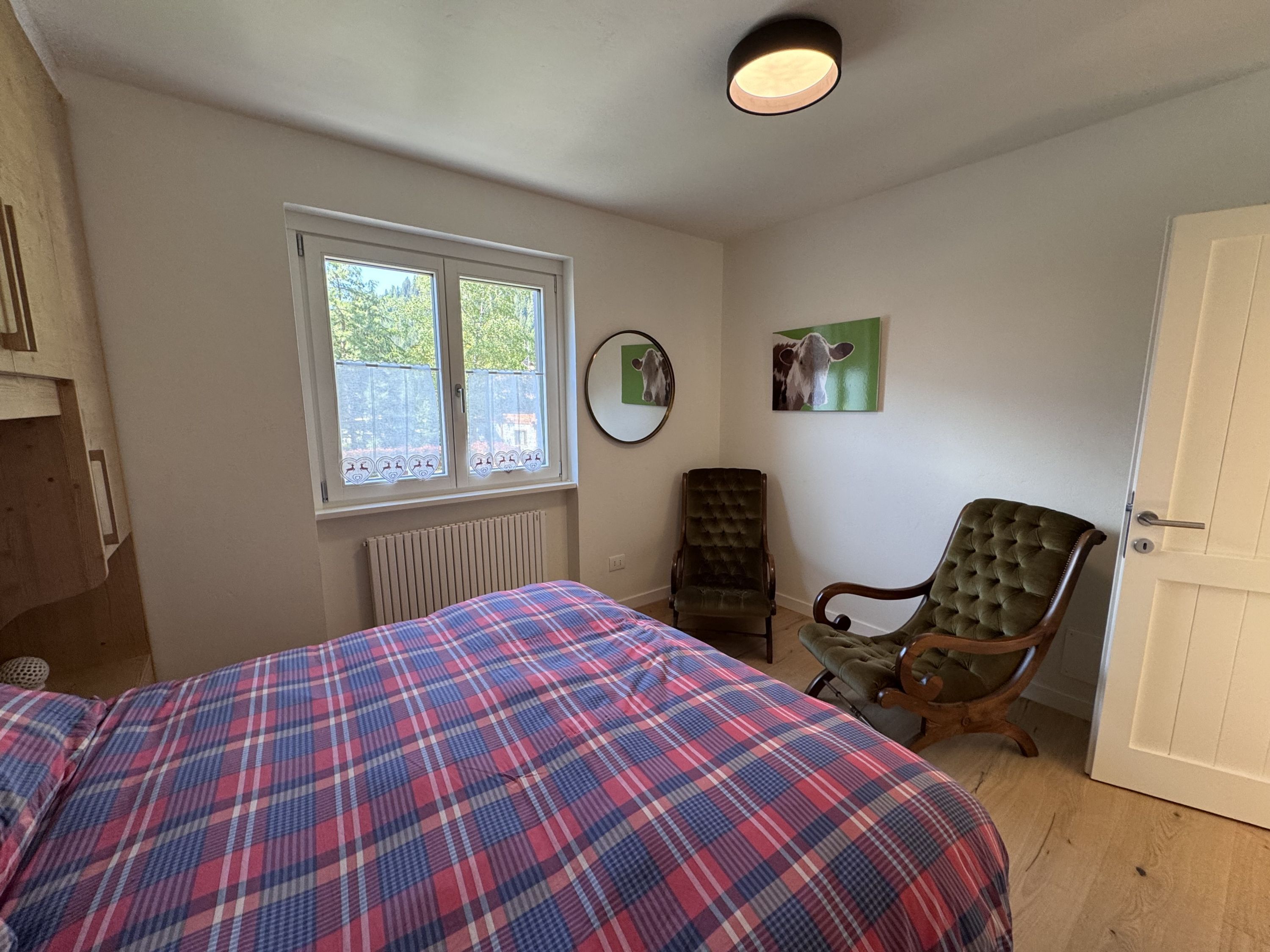A bedroom with a plaid bedspread, a radiator, a window with a view, a mirror, and a door.