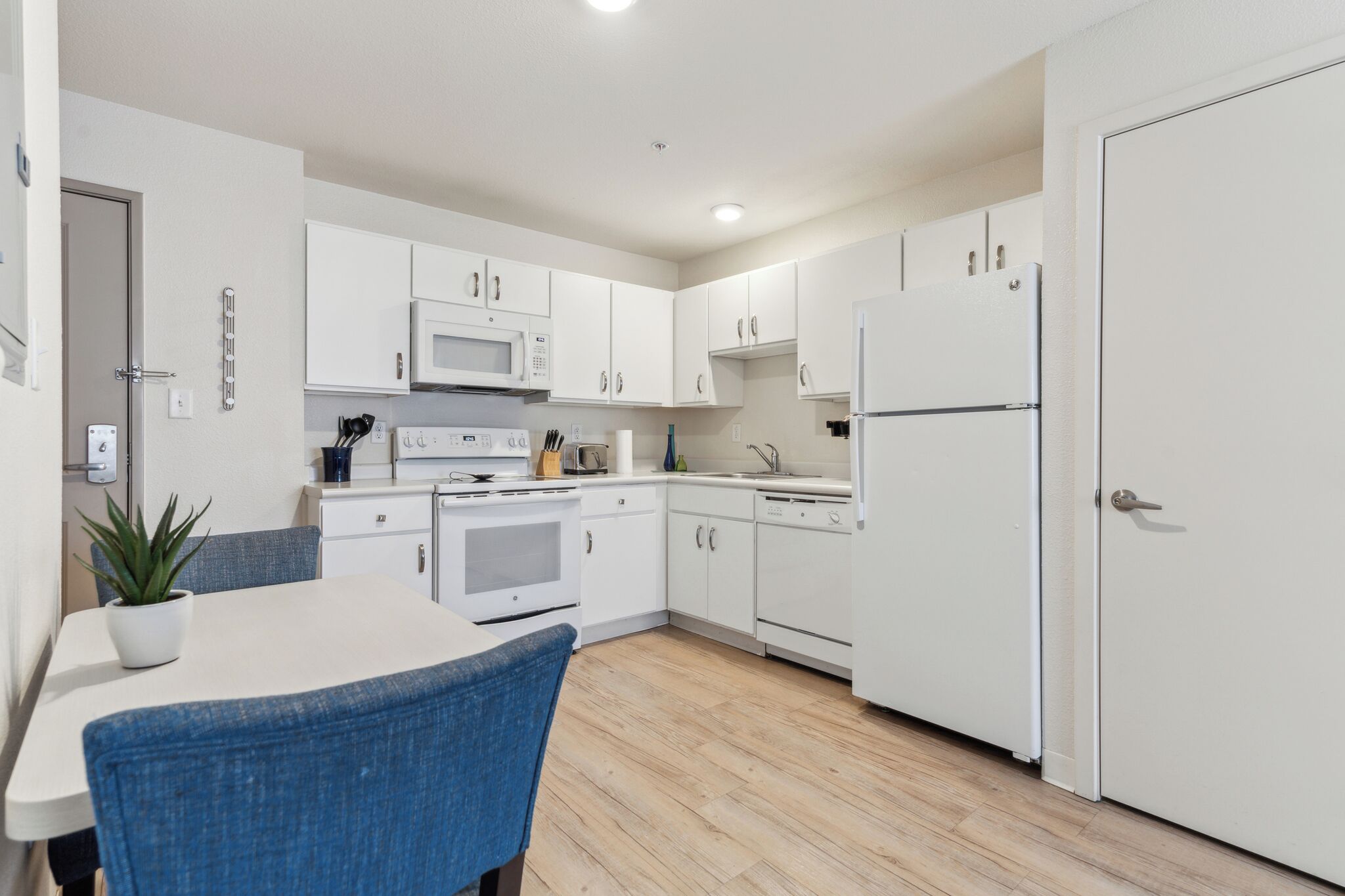 A modern kitchen with white cabinets, a microwave, stove, and refrigerator. There is a dining table with chairs and a potted plant.